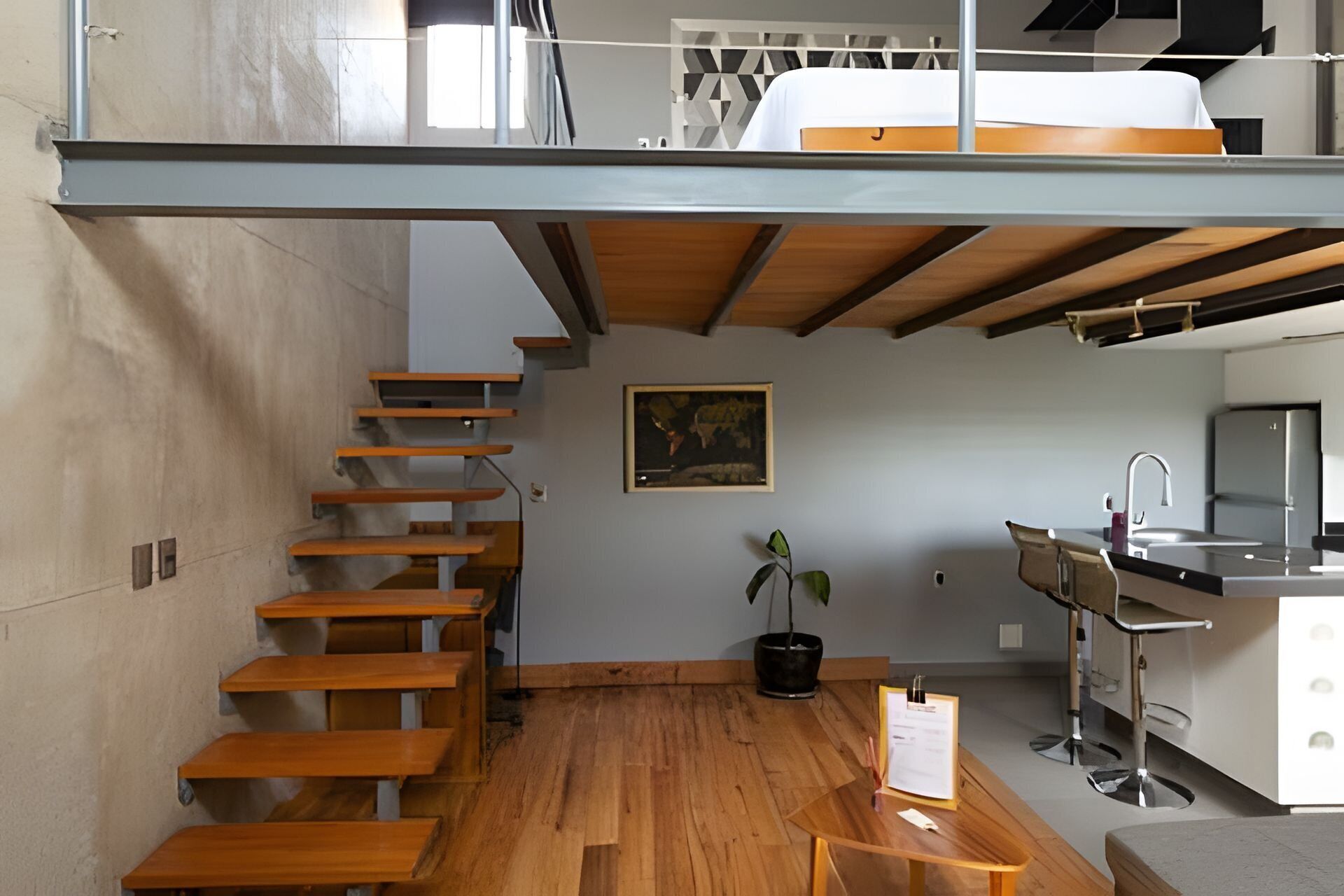 A modern loft-style apartment with a wooden staircase, a dining area, a kitchen with a sink, and a bed on the upper level.