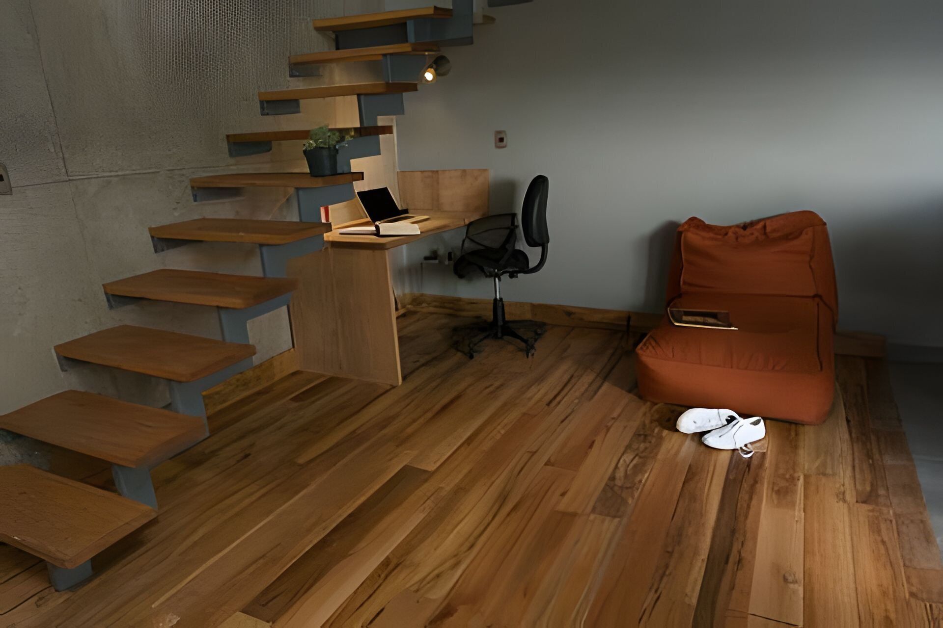 A modern room with a wooden staircase, a desk with a chair, a sofa, and a pair of shoes on the floor.