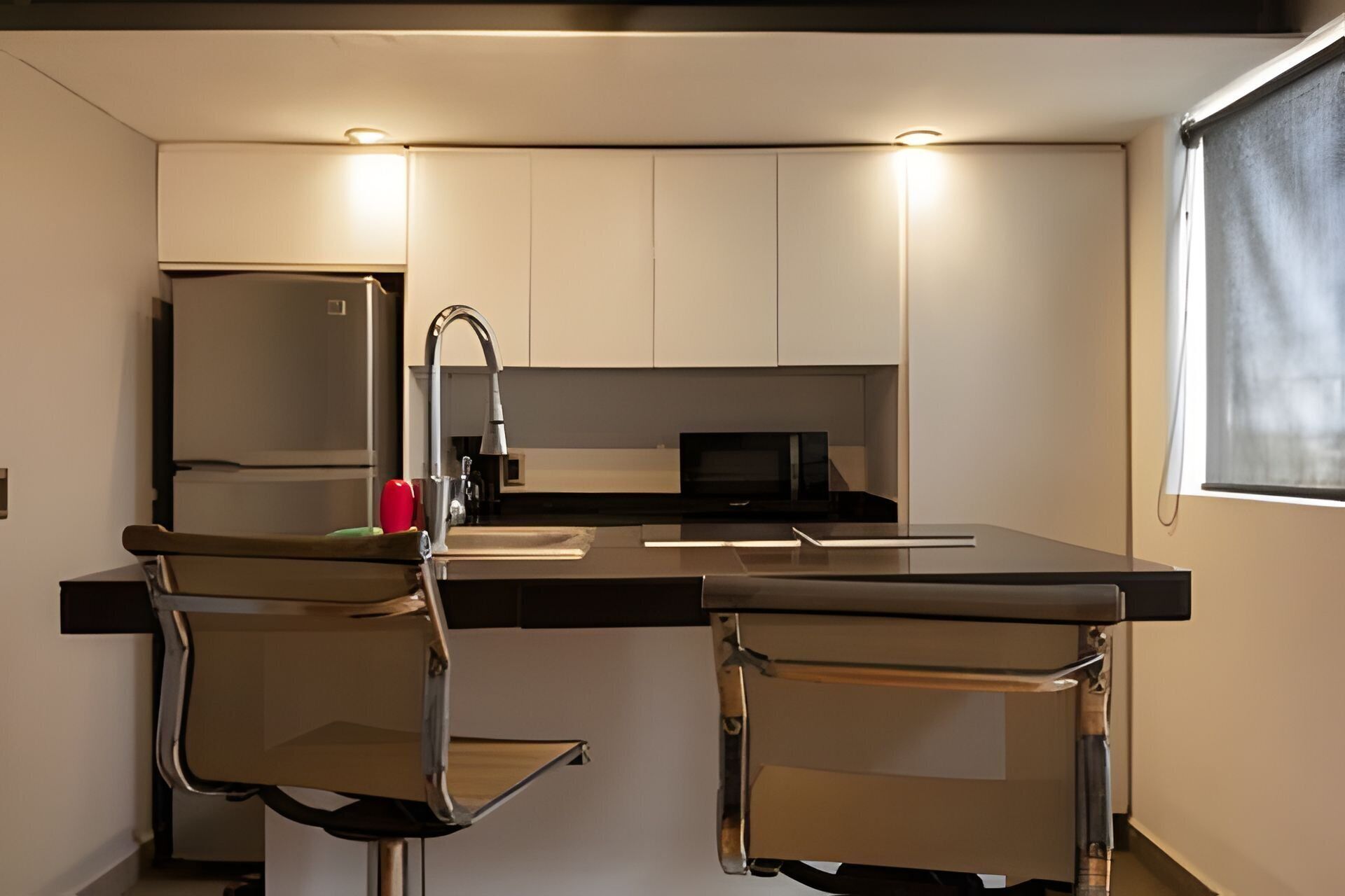 Una cocina moderna con una isla central, dos taburetes de bar, un microondas y un ventanal con persiana.
