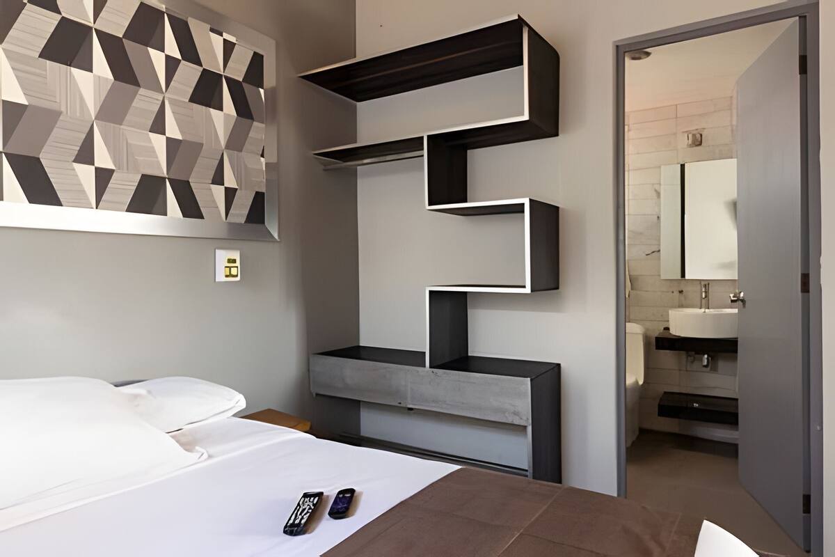 Quarto moderno com cama, arte de parede geométrica, estante embutida e banheiro visível através de uma porta aberta.