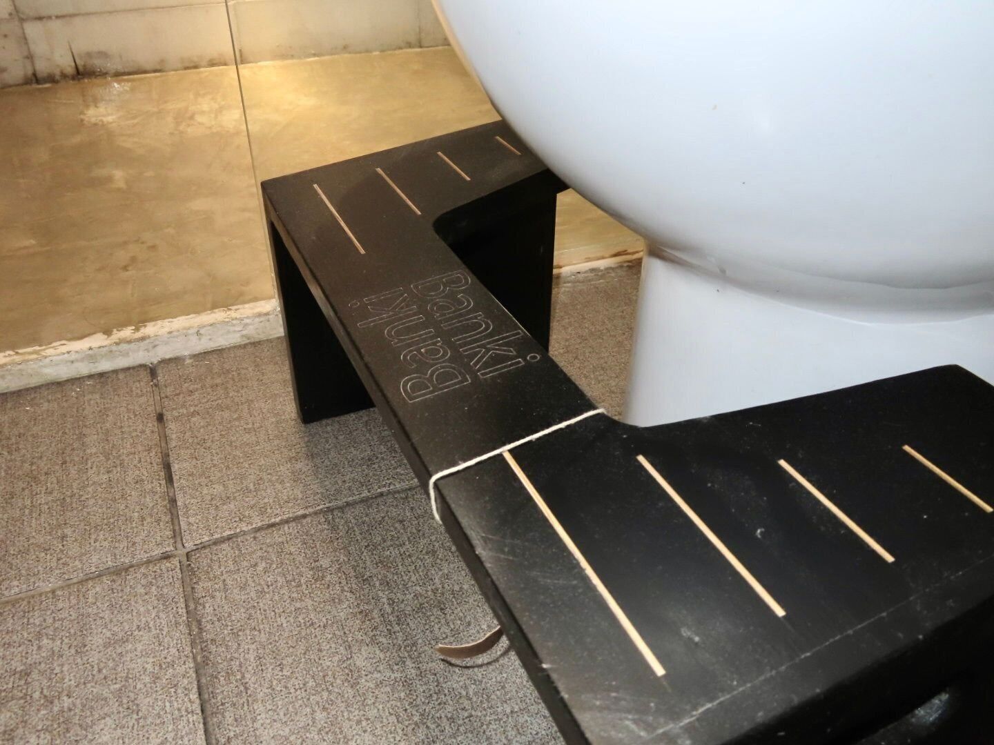 Et sort trin med teksten "Bank" og "Banka" på, placeret ved siden af et hvidt toilet i et badeværelse med flisegulv.