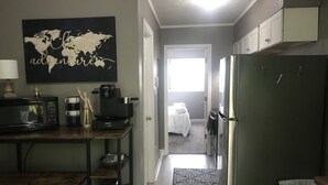 Una cocina compacta con microondas, cafetera y máquina para cápsulas de café Keurig. En la pared, un cuadro con un mapa del mundo, y una pequeña mesa de madera al frente.