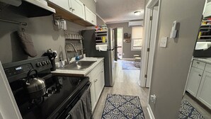 Una cocina con estufa, horno, pileta y armarios. Hay un tapete estampado en el piso y una puerta que da a otra habitación.