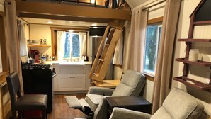 Un espace de vie compact comprenant un coin cuisine, un coin repas avec des chaises, une échelle en bois et une fenêtre avec des rideaux.