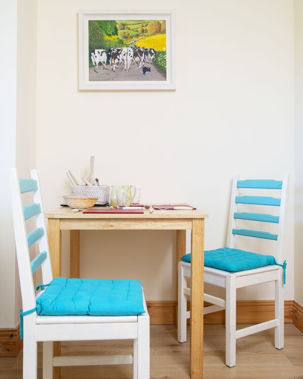 A room with a wooden table and two chairs with blue cushions. A framed painting of cows is on the wall.