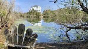 A wooden Adirondack chair with "ITS 5:00 SOMEWHERE" and "AFTERNOON DELIGHT" carved into it, positioned by a calm lake with a house in the background. - Soundside, 5 min walk to the beach, 6 private decks, shared pool and pickleball (Avon)