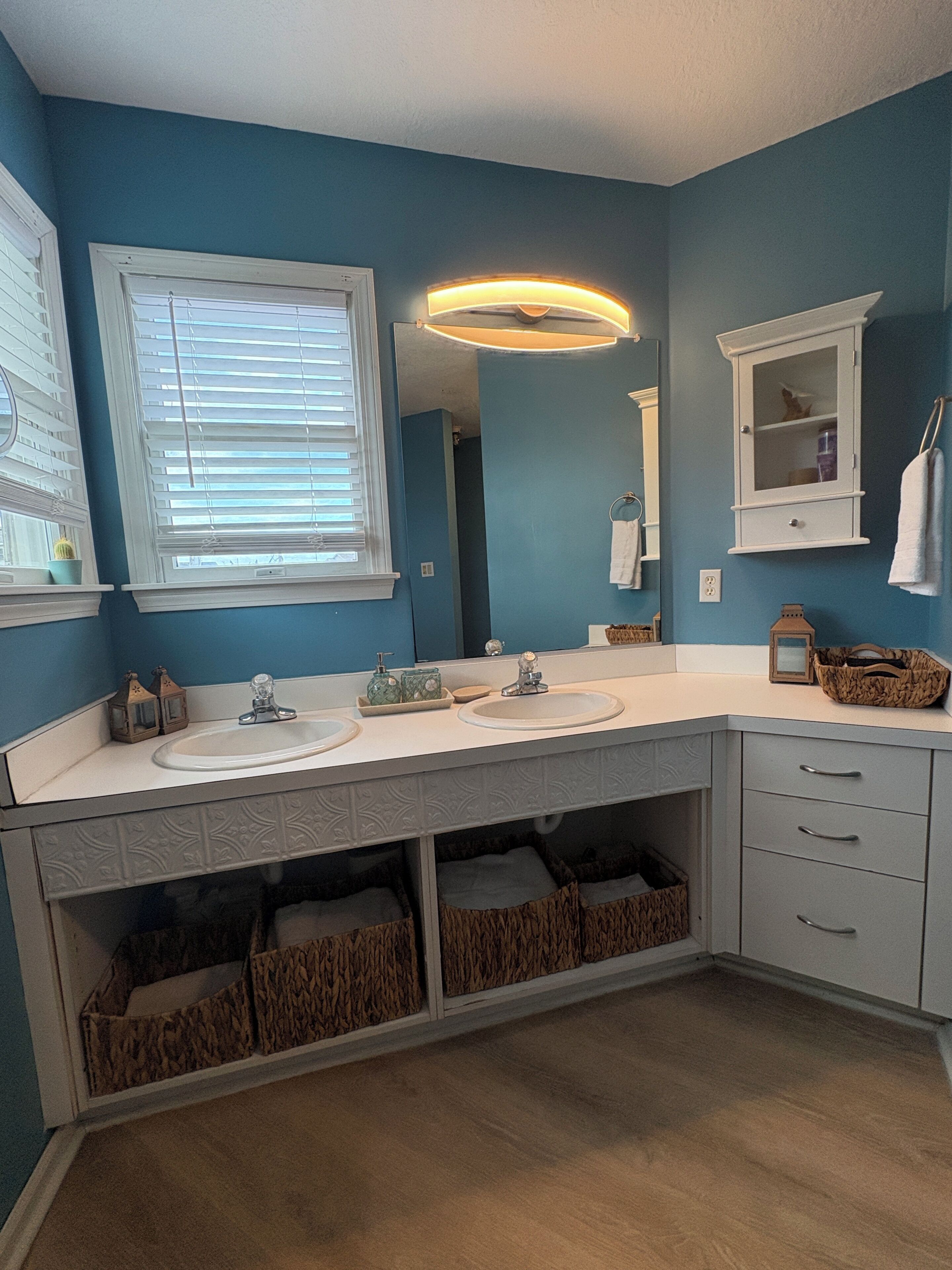 A bathroom with two sinks, a mirror, a window with blinds, and storage baskets under the sinks.