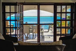 A room with a view of the ocean through a glass door, a dining area with chairs and a table, and a wicker sofa.