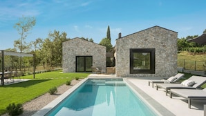 A modern outdoor pool area with two stone buildings, one with a glass door and the other with a large window, surrounded by a well-maintained lawn and a clear blue sky. - Villa Touch of Nature with Pool (Motovun)