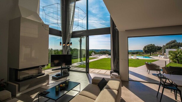 Ein modernes Wohnzimmer mit großem Fernseher, bequemen Sitzmöglichkeiten und Blick auf Pool und Garten.