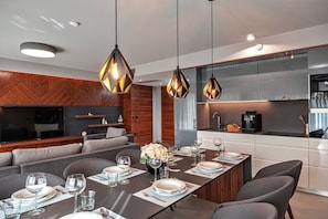A modern dining area with a long table set for six, a built-in kitchen with stainless steel appliances, and a cozy seating area with a flat-screen TV.