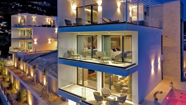 Un hotel moderno de varios pisos con fachada de vidrio, asientos al aire libre y montañas al fondo.
