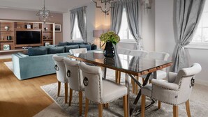 Una sala moderna con una mesa de comedor de vidrio rodeada por seis sillas tapizadas con anillos metálicos, un sofá azul, un televisor montado en un mueble de madera y una araña.