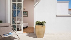 Un balcón bañado por el sol con una silla acolchada, una planta en maceta y una cama con dosel blanco.