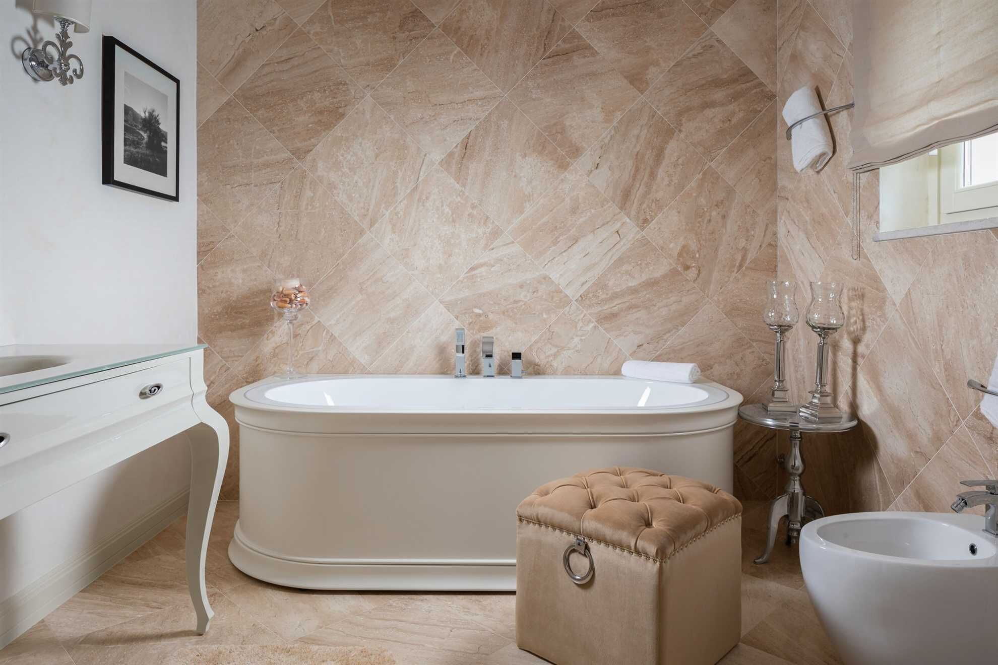 A modern bathroom with a freestanding bathtub, a vanity with a glass top, a beige ottoman with a ring handle, and a white sink.