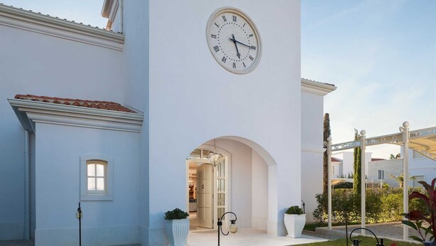 Un edificio blanco con un gran reloj en la fachada, un acceso cubierto y un camino de ladrillo que conduce a la entrada.