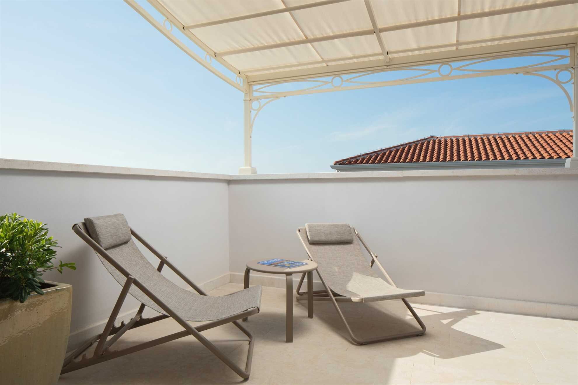 Eine Dachterrasse mit zwei Liegestühlen, einem kleinen Tisch, einer Topfpflanze und einer weißen Pergola.