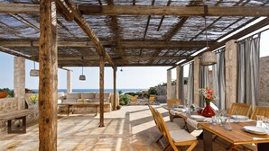 An outdoor patio with stone flooring, wooden furniture, and a view of the sea.