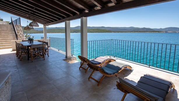 A covered outdoor area with a table and chairs, two sun loungers, and a view of a body of water with mountains in the background. - Villa Simpleza - Six Bedroom Villa with Private Pool and Jacuzzi (Klek)