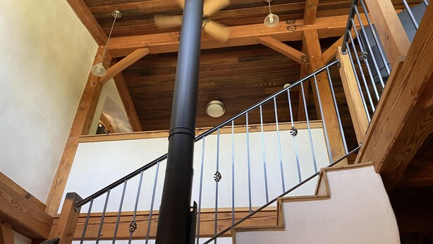 Un escalier en bois, doté d’un poteau central noir et de rampes en métal, mène à un niveau supérieur où l’on trouve des poutres en bois et un ventilateur de plafond.