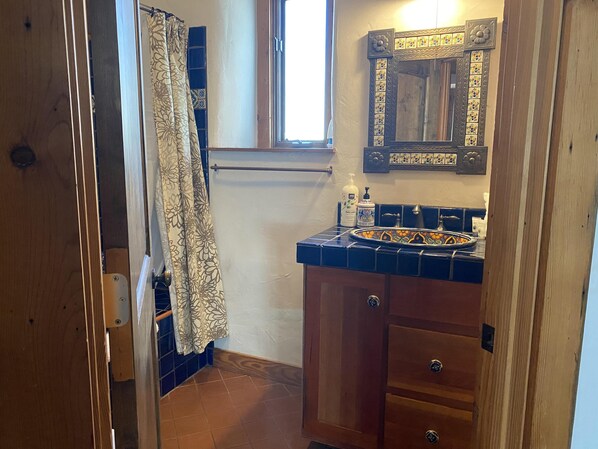 Une salle de bain avec un meuble en bois, un lavabo agrémenté d’un pare-feu en mosaïque décorative, un miroir avec un cadre floral et une fenêtre avec un rideau.
