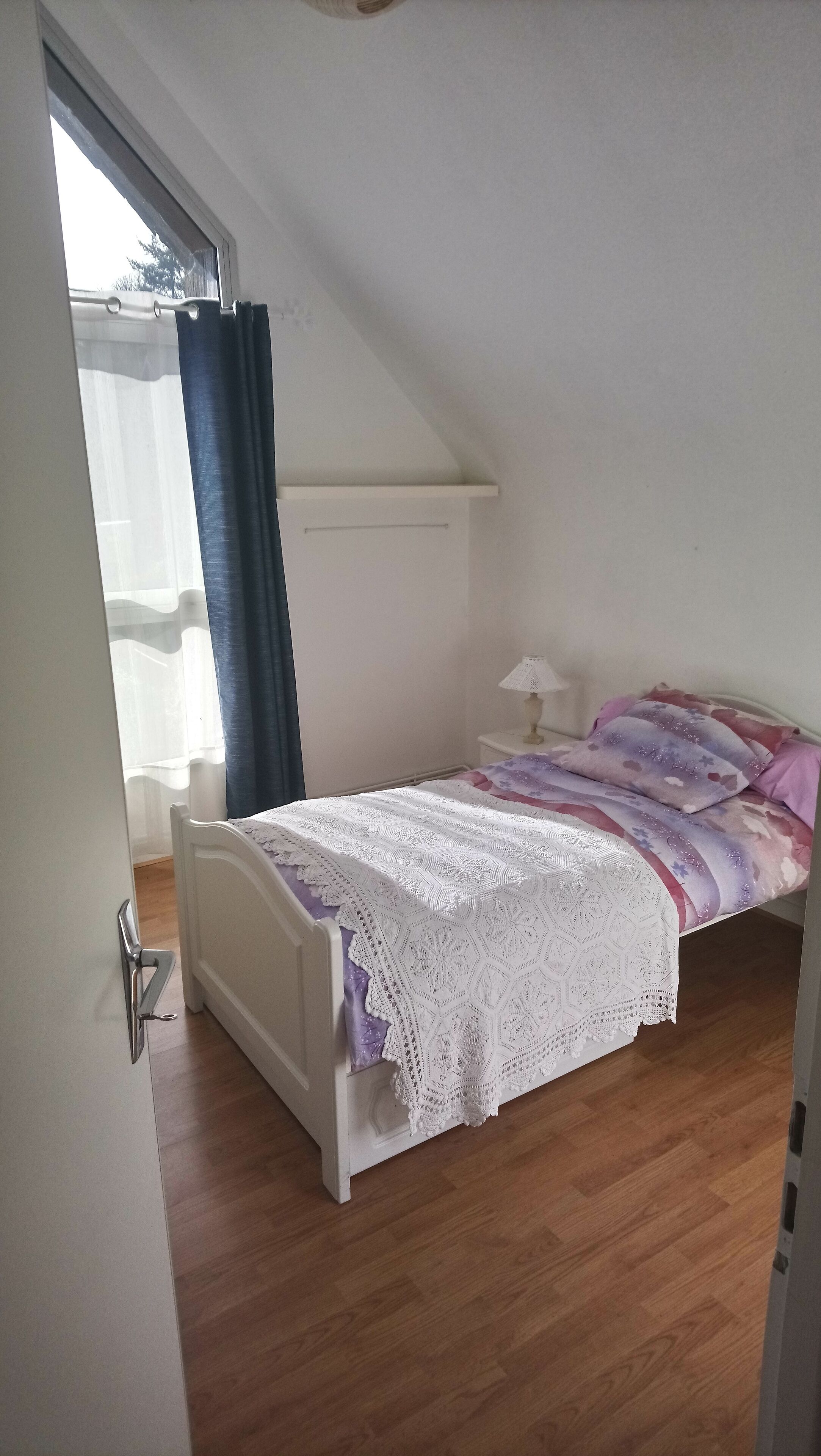 Ein Schlafzimmer mit schrägem Dach, einem Bett mit weißer Spitze, einem Nachttisch mit Lampe und einem Fenster mit Vorhängen.