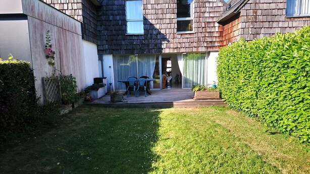 Un jardin à l’arrière d’une maison, avec une terrasse, une petite table et des chaises, ainsi qu’une haie.