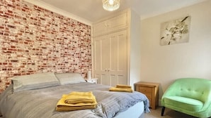 Una camera da letto con parete in mattoni, un letto con copripiumino a righe grigio e bianco, una poltrona verde e un comodino in legno.