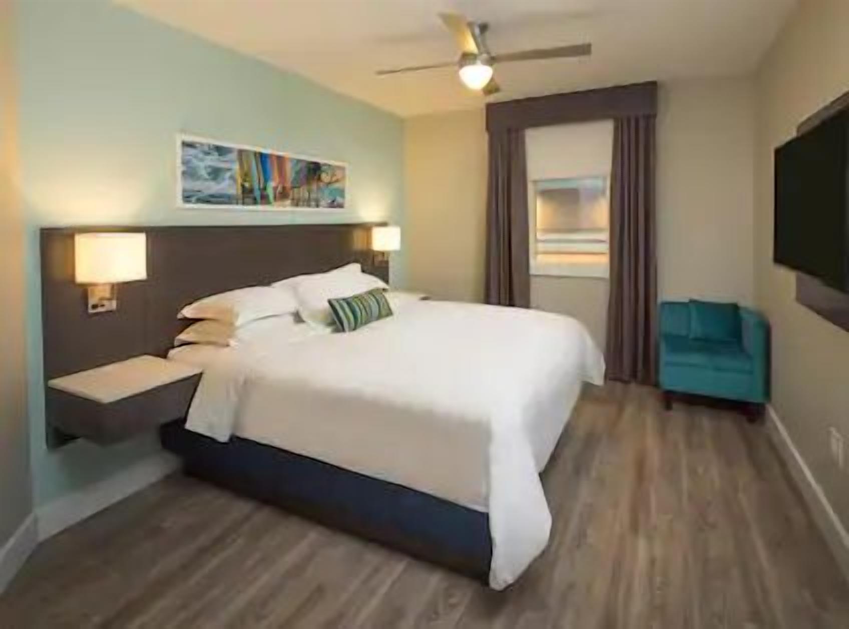 Una habitació d'hotel amb un llit, una taula de nit, un televisor penjat a la paret, un ventilador de sostre i una finestra amb cortines.