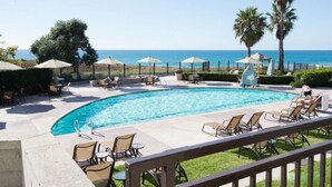 Area piscina con lettini e ombrelloni, vista sul mare e un edificio con una palma.