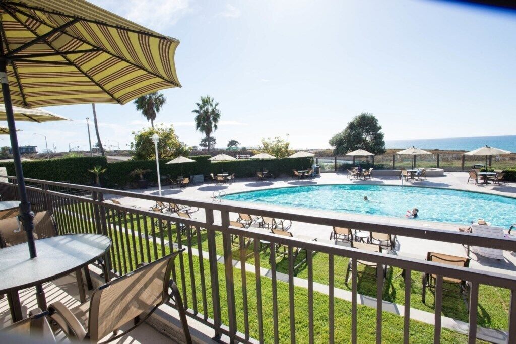 Un espace piscine avec des chaises longues et des parasols, une vue sur l’océan et une terrasse entourée d’une clôture.