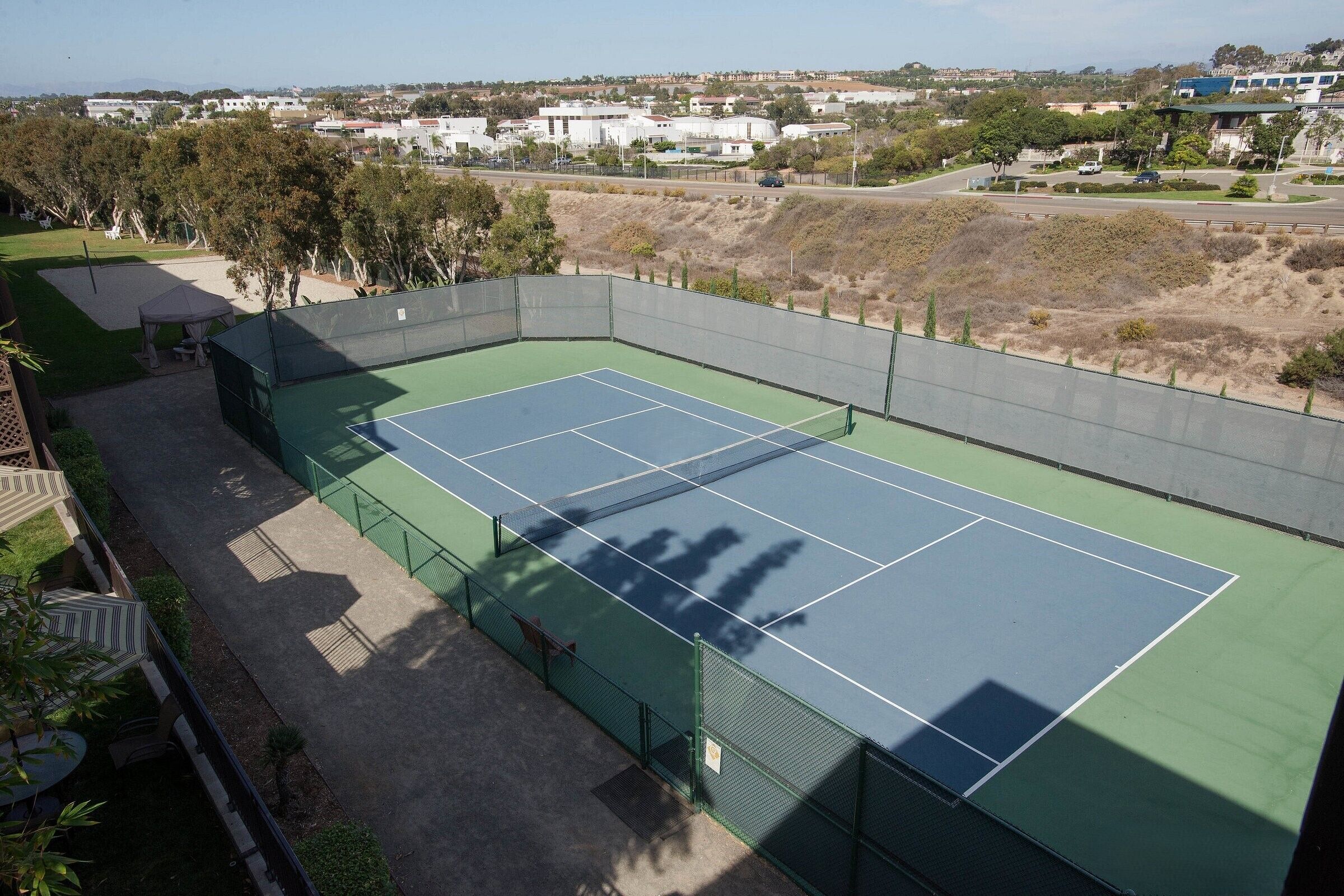 Un court de tennis doté d’une surface verte, entouré d’une clôture, et offrant une vue sur un quartier résidentiel en arrière-plan.