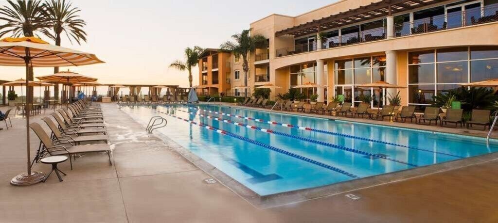 A swimming pool with lounge chairs and umbrellas, surrounded by a building with balconies.