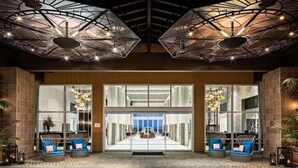 Un vestíbulo de hotel de diseño moderno, con entrada de vidrio, zona de estar con sillas de mimbre y una estructura de techo singular.