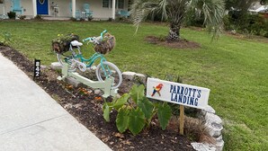 Un panneau décoré de motifs de perroquets, un vélo turquoise et une maison avec une porte bleue et des chaises sur le porche.