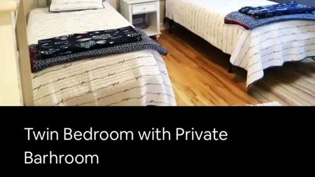 Une chambre double avec salle de bain privée, comprenant deux lits avec du linge de lit à rayures, une petite table de chevet avec une lampe et une guirlande décorative accrochée au mur.