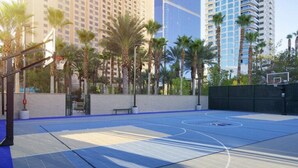 An outdoor basketball court with a blue surface, surrounded by palm trees and tall buildings in the background.