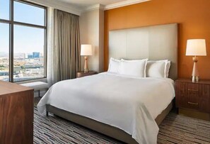 A hotel room with a large bed, two bedside tables with lamps, a view of the city through a large window, and a wooden desk. - Las Vegas | Pool & Strip Access | 3BR Sleeps 8 (Las Vegas)
