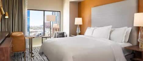 A hotel room with a large bed, a desk, a chair, and a view of a cityscape through a large window.
