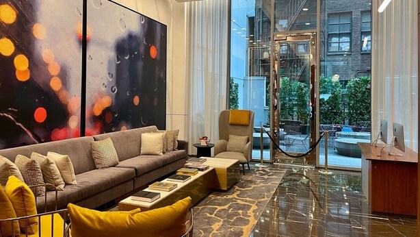 A modern living room with a sofa, armchair, and coffee table, large abstract paintings, and a view of the city through large windows. - NYC | 5th Ave Location | Central Park | 1BR Penthouse (New York)