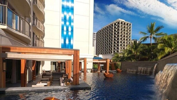 A hotel pool area with a waterfall, lounge seating, and a modern building in the background. - Waikiki | Friends Hawaii Beach Trip | Pool + Beach (Honolulu)