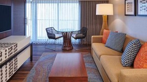 A hotel room with a sofa, a coffee table, a TV, a dining table with chairs, and a large window with curtains. - Waikiki | Friends Hawaii Beach Trip | Pool + Beach (Honolulu)