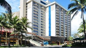 A multi-story hotel with balconies, a blue vertical feature, and palm trees. - Waikiki | Friends Hawaii Beach Trip | Pool + Beach (Honolulu)