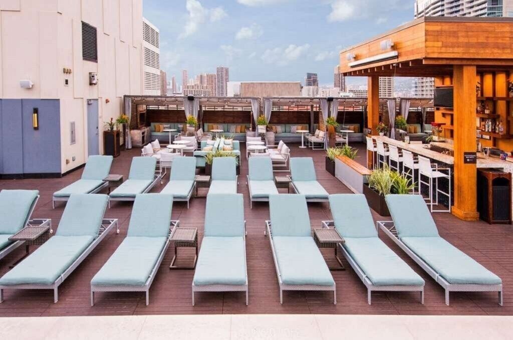 A rooftop lounge area with sun loungers, a bar, and a city view.