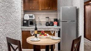 A compact kitchen with wooden cabinets, a refrigerator, a microwave, and a coffee maker. There is a dining table with chairs and a bowl of fruit.