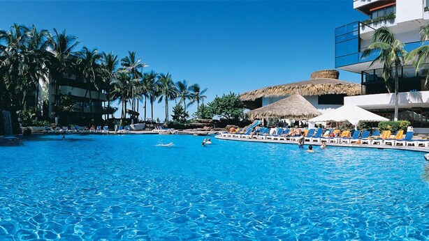 A large swimming pool with lounge chairs and palm trees, surrounded by buildings.