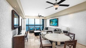 A spacious room with a large window offering a view of the sea, a ceiling fan, a flat-screen TV, a round dining table with chairs, and a sofa with cushions.