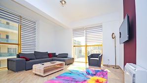 A modern living room with a grey sofa, a colorful rug, and a large window with blinds. - Orange Lux Apartments (Budva)