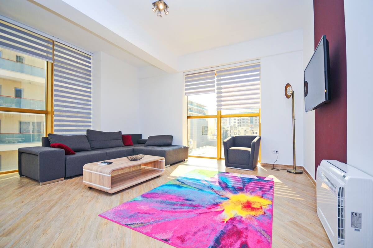 A modern living room with a grey sofa, a colorful rug, and a large window with blinds.
