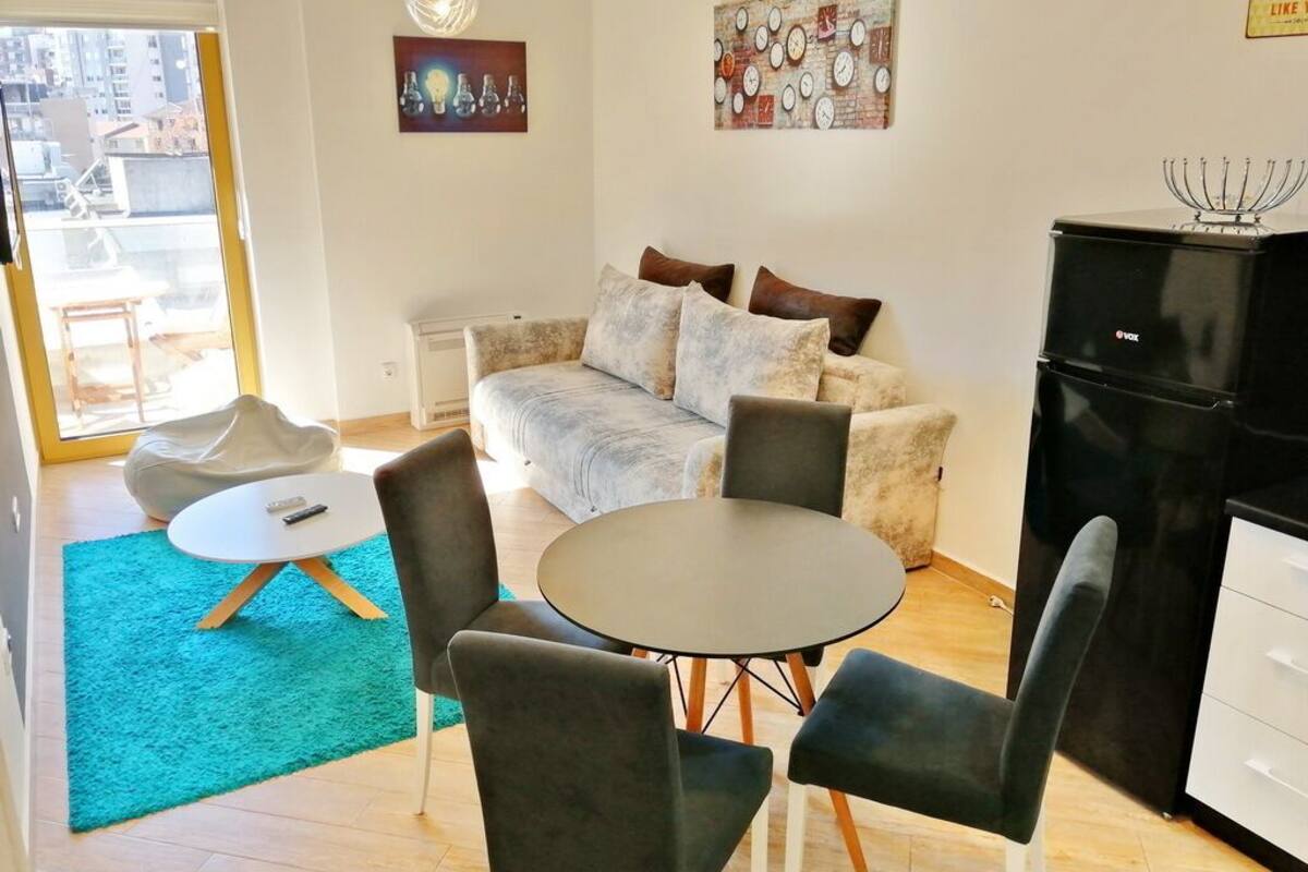 A living room with a sofa, a round table, and chairs. There is a black cabinet and a window with a view of buildings outside.